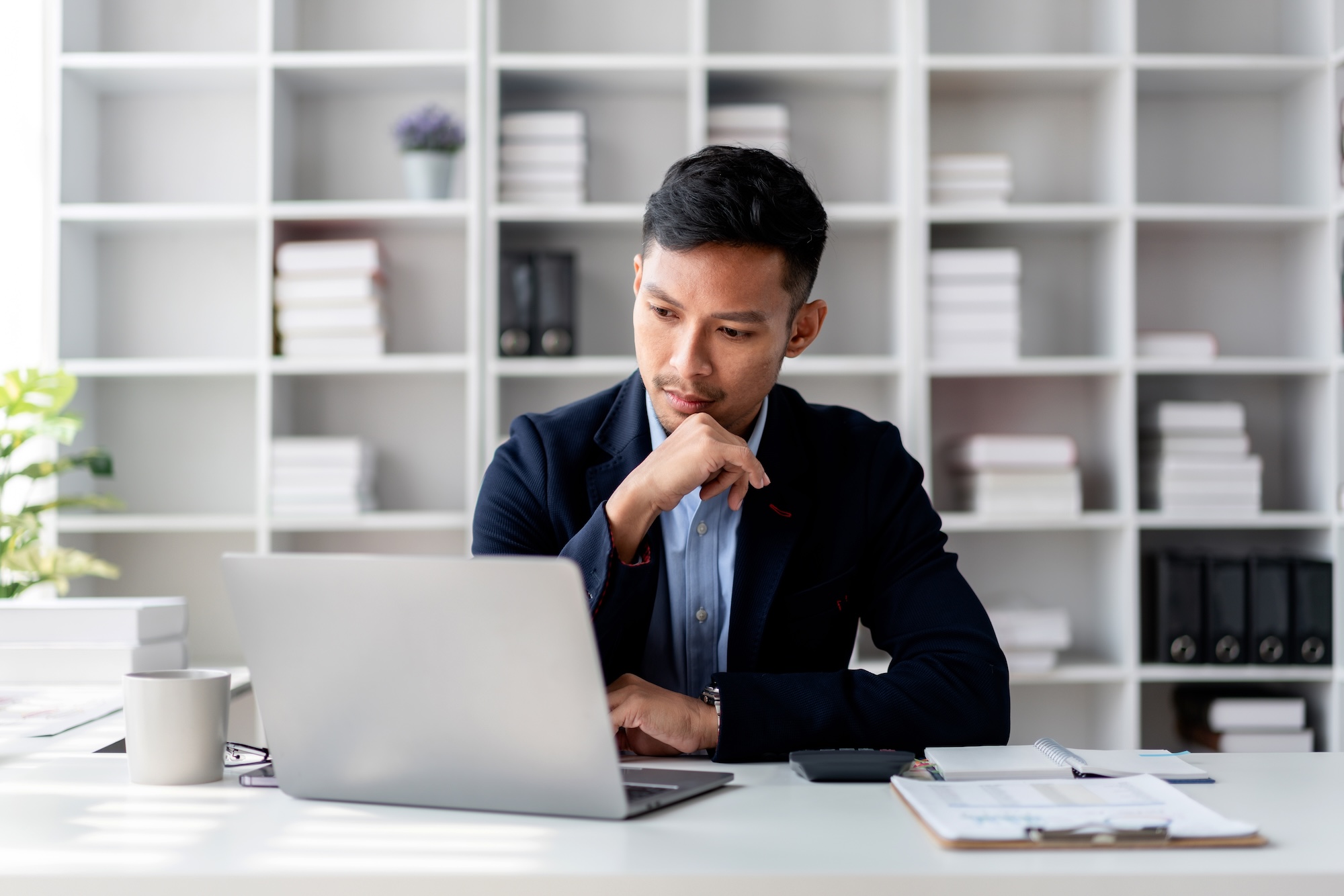 Adult Asian businessman in suit working on laptop in bright office. He appears to be concentrating and planning business strategy using computer and internet for his work.